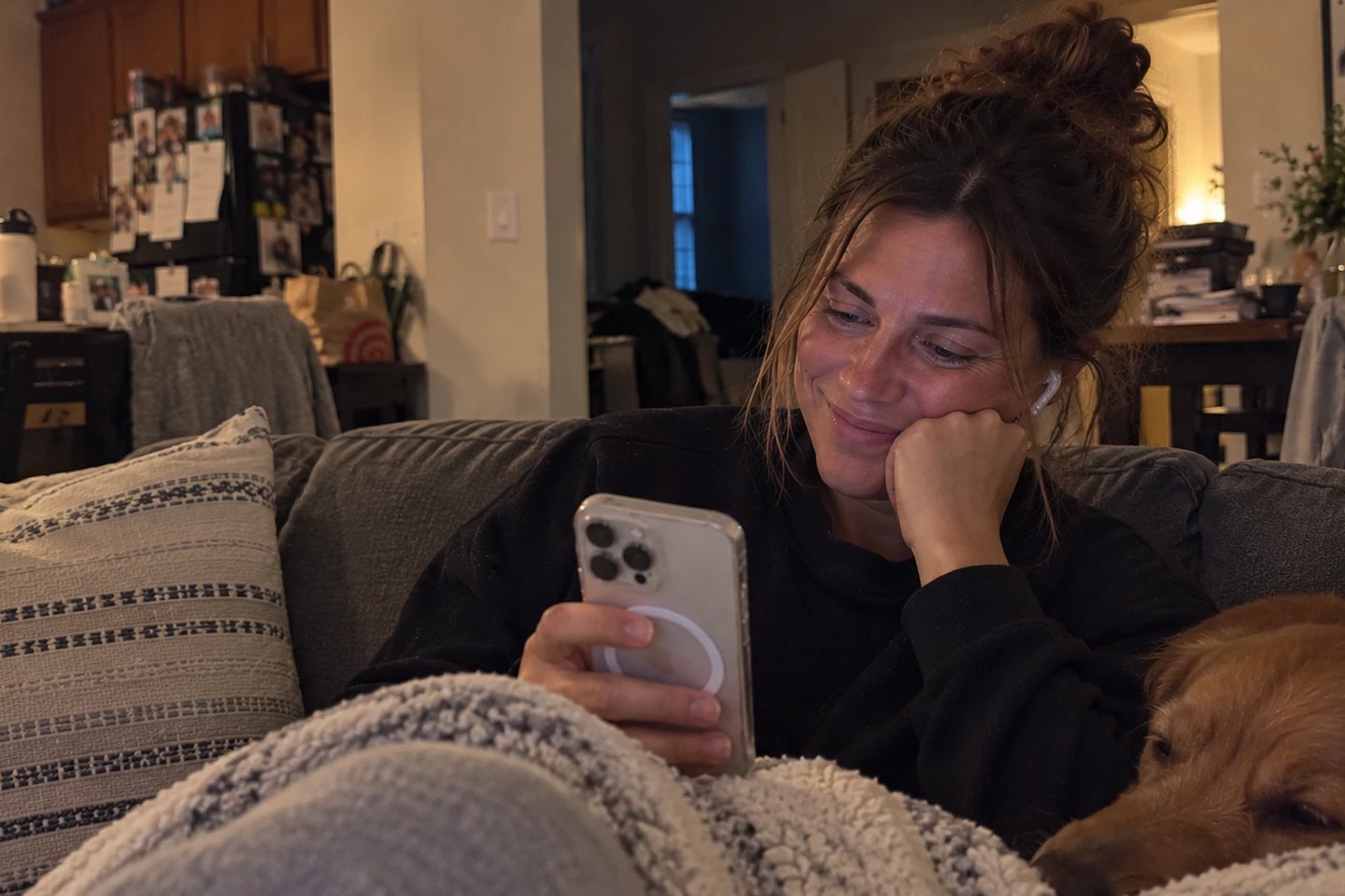 A woman curled up on her sofa with her dog, smiling and tearful as she listens to a song on her phone.