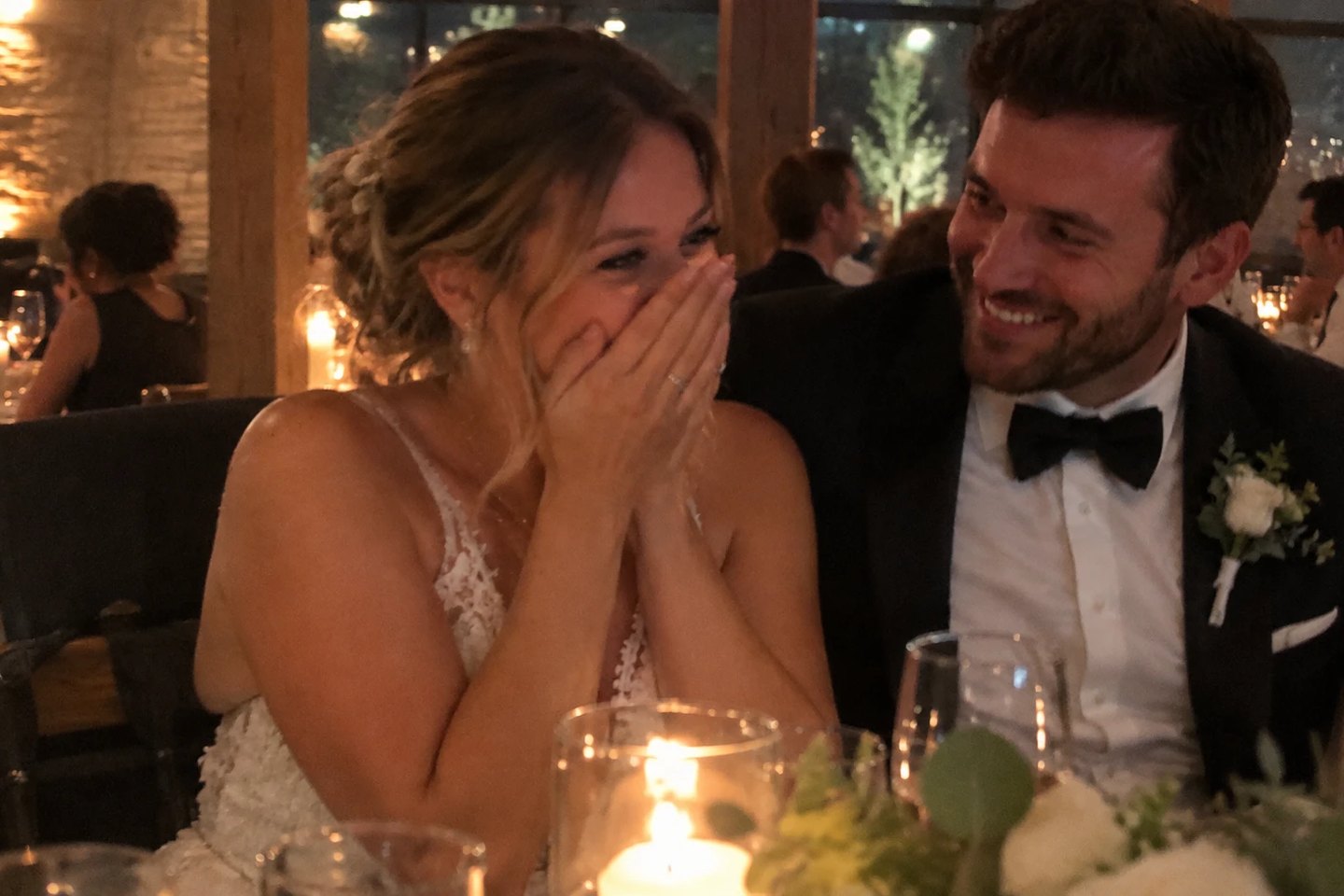 A bride laughing behind her hands as the groom smiles at her during their candlelit wedding reception.